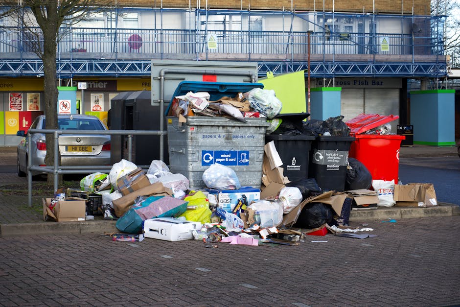 A pile of various waste materials, including cardboard boxes, paper bags, and plastic packaging, is situated against a brick wall on a pavement or ground surface. The boxes are in different states of 