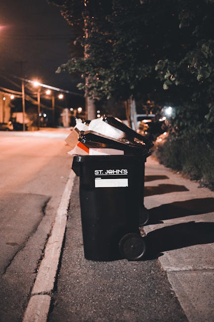 Two large black plastic rubbish bags filled with waste are positioned on the pavement near the edge of a street, leaning against a wooden fence with vertical slats. The bags are slightly crumpled and 