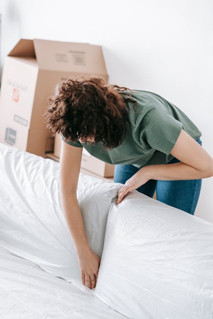 A woman with curly brown hair, dressed in a green short-sleeved top and blue jeans, is seen preparing a white mattress for disposal or removal indoors. She is leaning forward, carefully rolling or fol
