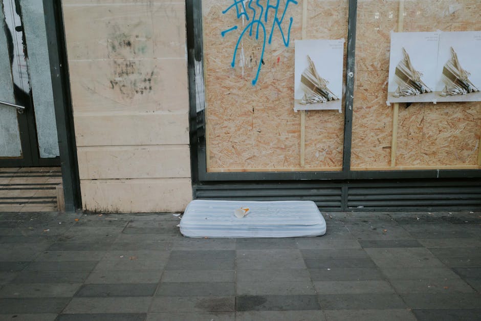 A woman with curly brown hair, dressed in a green short-sleeved top and blue jeans, is seen preparing a white mattress for disposal or removal indoors. She is leaning forward, carefully rolling or fol