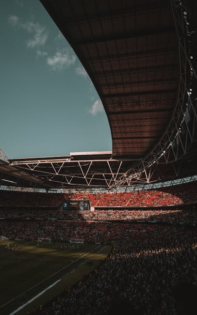 A wide-angle view of Wembley Stadium's interior during an event, showing the curved metallic roof structure supported by angular trusses that extend over the seating area. The seating is filled with s