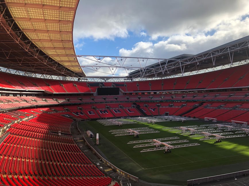 A wide-angle view of Wembley Stadium's interior during an event, showing the curved metallic roof structure supported by angular trusses that extend over the seating area. The seating is filled with s
